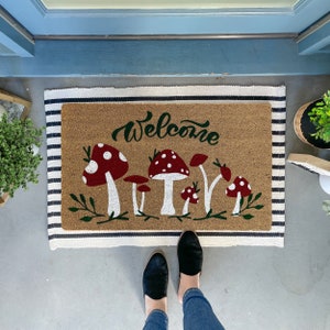 May include: A welcome mat with a brown background and black and white stripes. The mat has the word "Welcome" written in cursive and a design of red and white mushrooms with green stems and leaves.