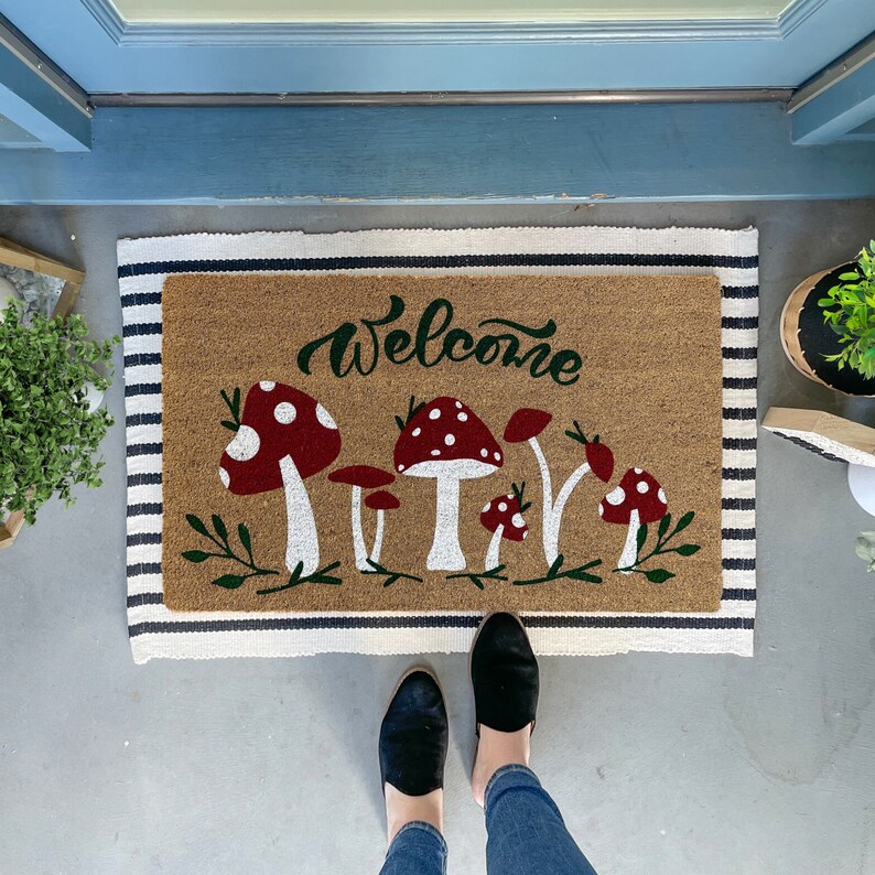 May include: A welcome mat with a brown background and black and white stripes. The mat has the word "Welcome" written in cursive and a design of red and white mushrooms with green stems and leaves.