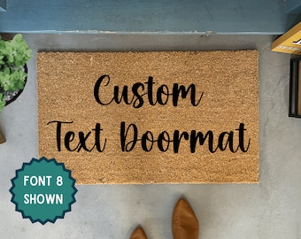 May include: A rectangular coir doormat with the words "Custom Text Doormat" in black script. A teal badge on the left reads "FONT 8 SHOWN". The doormat is on a gray surface, with a blue door frame and a small plant visible.