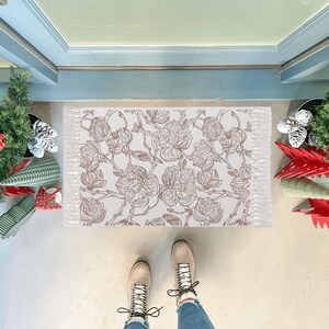 May include: A rectangular doormat featuring a repeating brown line-art floral pattern on a white background. The mat has a textured surface and short white fringe detailing along the two shorter sides, positioned on a light-colored floor.