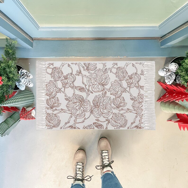 May include: A rectangular doormat featuring a repeating brown line-art floral pattern on a white background. The mat has a textured surface and short white fringe detailing along the two shorter sides, positioned on a light-colored floor.