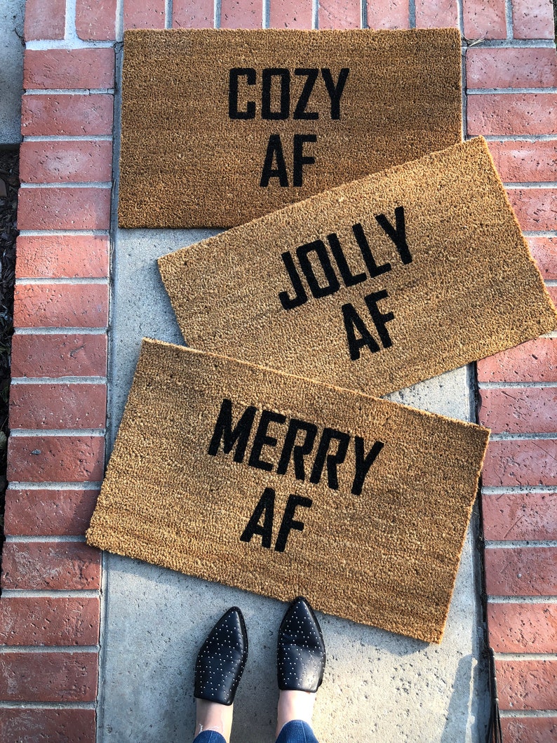 May include: Three brown doormats with the text "Cozy AF", "Jolly AF", and "Merry AF" in black lettering.
