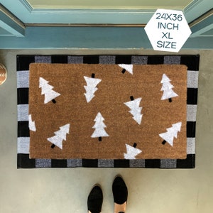 May include: A brown doormat with a white checkered border and a pattern of white Christmas trees. The doormat is 24x36 inches and is labeled "XL Size".