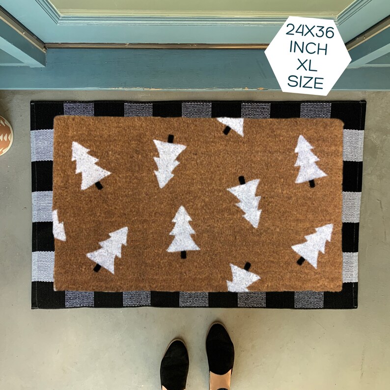 May include: A brown doormat with a white checkered border and a pattern of white Christmas trees. The doormat is 24x36 inches and is labeled "XL Size".