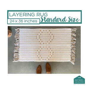 May include: A rectangular off-white layering rug measuring 24 x 36 inches with tan woven geometric patterns and fringed edges on the short sides. The text LAYERING RUG Standard Size 24 x 36 inches is displayed above the rug, and a logo is below.
