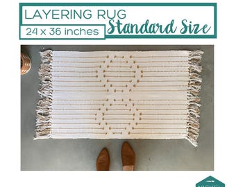 May include: A rectangular off-white layering rug measuring 24 x 36 inches with tan woven geometric patterns and fringed edges on the short sides. The text LAYERING RUG Standard Size 24 x 36 inches is displayed above the rug, and a logo is below.