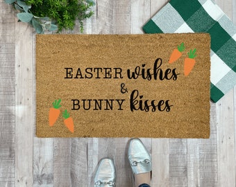 May include: A brown coir doormat with the text "Easter wishes & bunny kisses" in black lettering. There are orange carrot illustrations on the mat.
