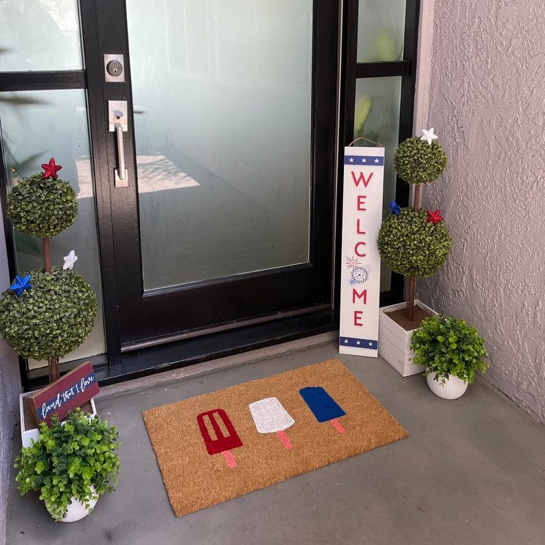 May include: A welcome sign with red, white and blue stars and fireworks, with the word "WELCOME" in bold letters. The sign is hanging on a door with a doormat featuring lolly graphics. Two potted plants with red, white and blue accents are on either side of the door.
