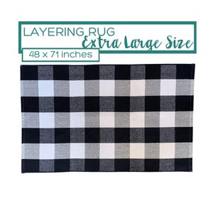 Może przedstawiać: Prostokątny dywanik w czarno-biało-szarą kratę. Tekst LAYERING RUG Extra Large Size 48 x 71 inches znajduje się nad dywanikiem w ramce z turkusową obwódką, wskazując wymiary przedmiotu podane w calach.