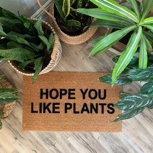 K&ouml;nnte beinhalten: Eine braune Kokosmatte mit dem Text "HOPE YOU LIKE PLANTS" in schwarzen Buchstaben. Die Matte ist von Topfpflanzen umgeben.