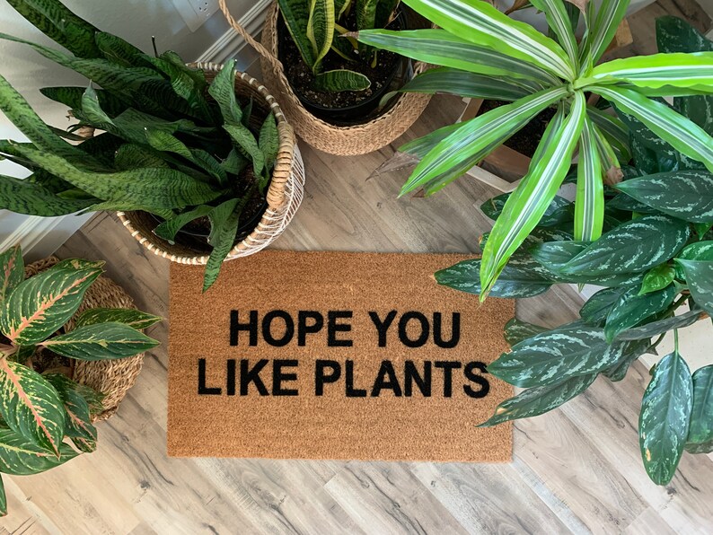 K&ouml;nnte beinhalten: Eine braune Kokosmatte mit dem Text "HOPE YOU LIKE PLANTS" in schwarzen Buchstaben. Die Matte ist von Topfpflanzen umgeben.