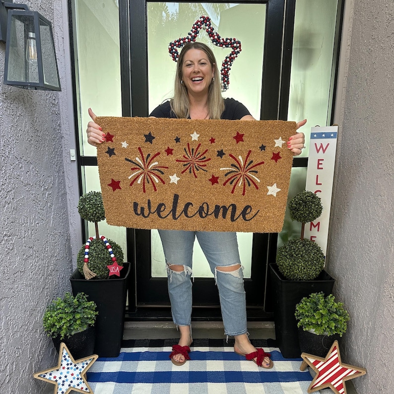 Patriotic summer doormat for front porch with red, white and blue fireworks design and bold script welcome across the bottom. this outdoor doormat is being held by a woman on a front porch decorated for the4th of july.