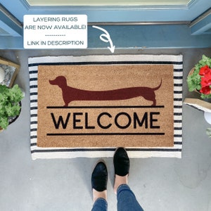 May include: A brown coir doormat with a black and white striped border. The doormat features a silhouette of a dachshund and the word "WELCOME" in black lettering.