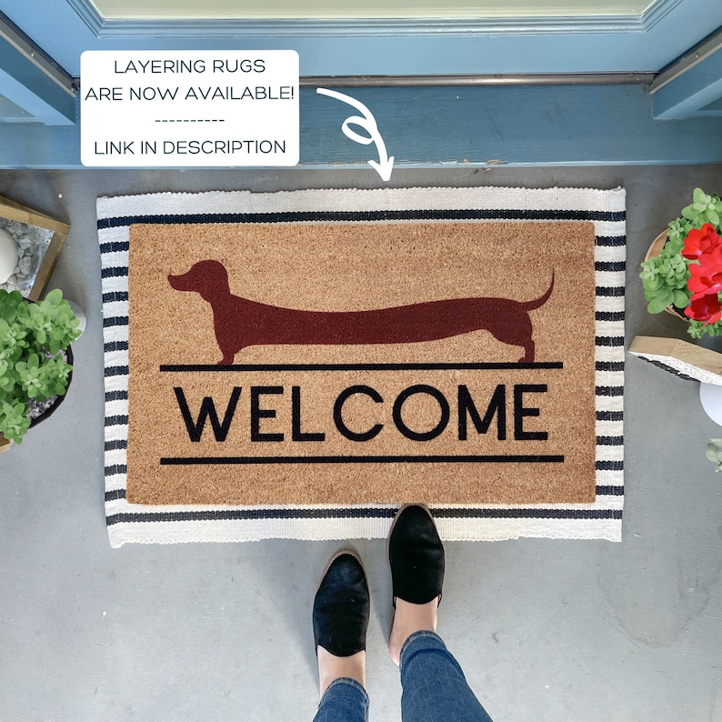 May include: A brown coir doormat with a black and white striped border. The doormat features a silhouette of a dachshund and the word "WELCOME" in black lettering.