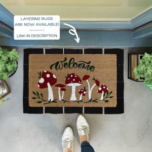 May include: A welcome mat with a brown coir background and black trim. The mat features a white "Welcome" inscription with green lettering and a row of five red and white mushrooms with green leaves.