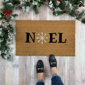 May include: A brown coir doormat with the word "NOEL" in black letters and a white snowflake in the center.