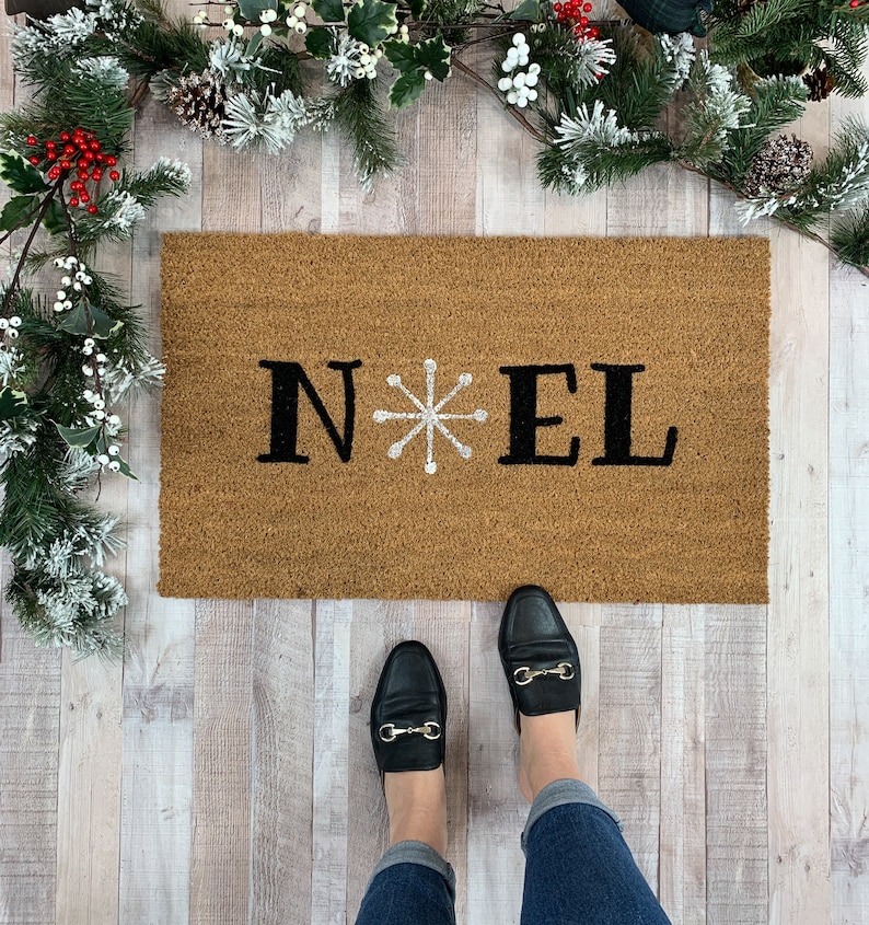May include: A brown coir doormat with the word "NOEL" in black letters and a white snowflake in the center.