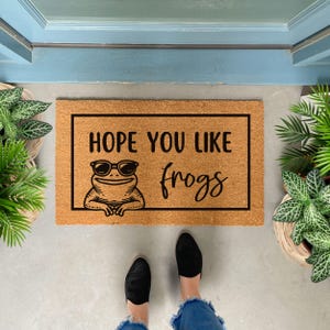 May include: A rectangular doormat made of natural coir fiber. The mat features a black border and the text "HOPE YOU LIKE frogs" with a cartoon frog wearing sunglasses. The mat is placed in front of a blue door.