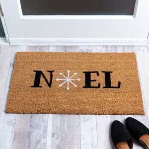 May include: A brown coir doormat with the word "NOEL" in black letters and a white snowflake in the center.