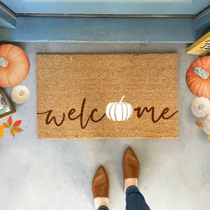 May include: A brown coir doormat with the word "welcome" written in brown script with a white pumpkin in the center of the word.