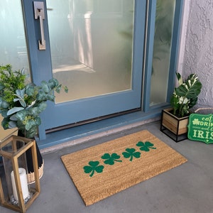 May include: A brown coir doormat with a row of five green shamrock designs. The doormat is placed in front of a blue door with a silver door handle. A wooden sign with the text "eat, drink and be Irish" is on the right side of the doormat.