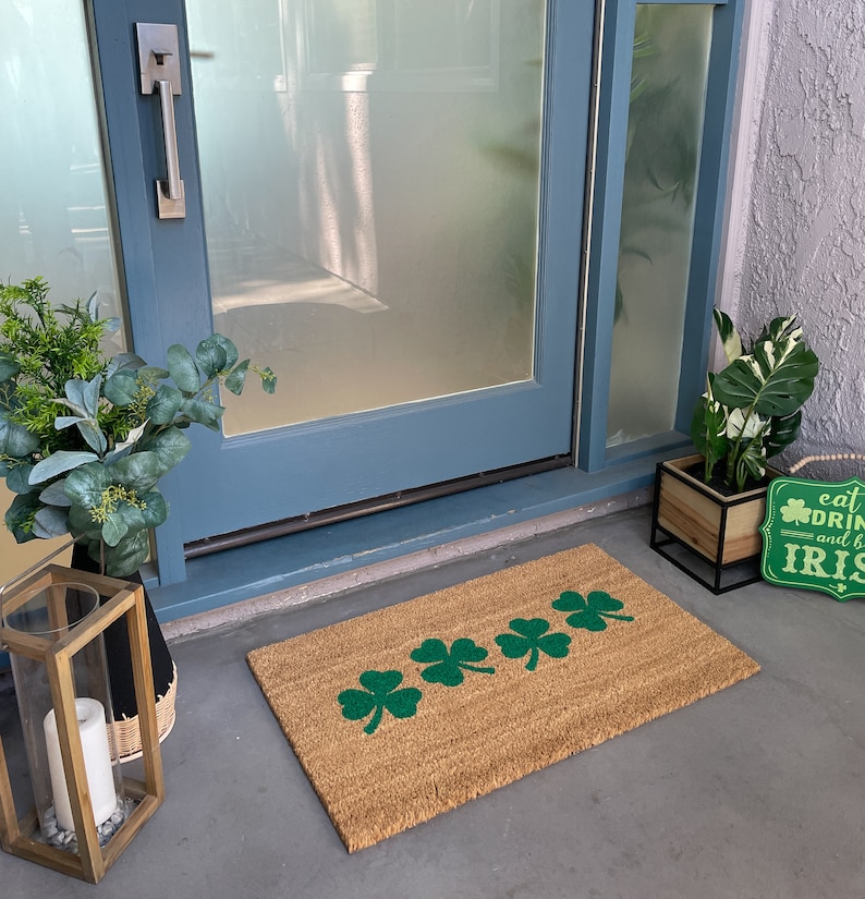 May include: A brown coir doormat with a row of five green shamrock designs. The doormat is placed in front of a blue door with a silver door handle. A wooden sign with the text "eat, drink and be Irish" is on the right side of the doormat.
