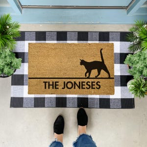 May include: A brown coir doormat with black and white chequered backing. The doormat has a black silhouette of a cat walking and the text "THE JONESES" printed on it.