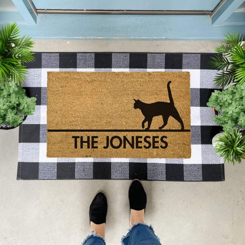 May include: A brown coir doormat with black and white chequered backing. The doormat has a black silhouette of a cat walking and the text "THE JONESES" printed on it.