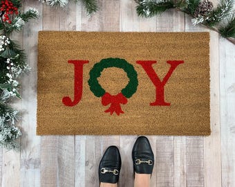 May include: A rectangular tan coir doormat featuring the word JOY in red, with a green wreath replacing the letter O and a red bow at the bottom of the wreath. The mat is positioned on a light-colored wooden floor surface.