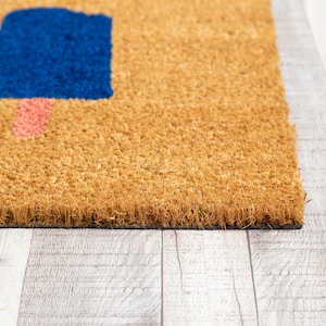 May include: A brown coir doormat with a blue and pink geometric design. The doormat has a black border and is sitting on a light wood floor.