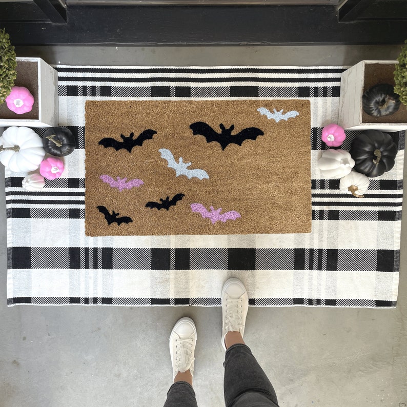 May include: A brown doormat with a black and white buffalo check pattern background. The doormat has a variety of bats in black, pink, and light blue colors.