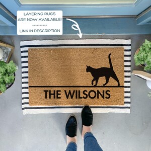 May include: A brown coir doormat with black stripes around the edges. The doormat has a black silhouette of a cat walking and the text "THE WILSONS" in black lettering.