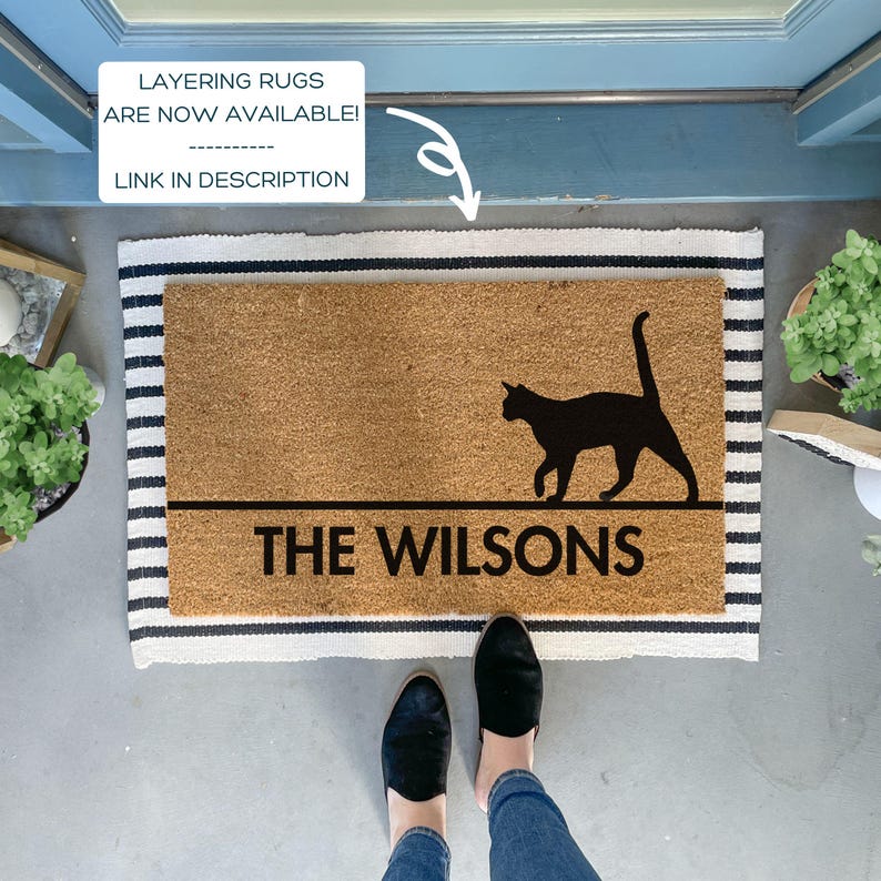 May include: A brown coir doormat with black stripes around the edges. The doormat has a black silhouette of a cat walking and the text "THE WILSONS" in black lettering.