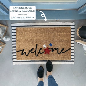 May include: A welcome mat with the word "welcome" in black script with a red, white, and blue star design. The mat is on a striped rug.