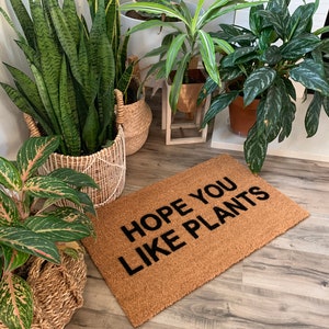 K&ouml;nnte beinhalten: Eine braune Kokosmatte mit dem Text "HOPE YOU LIKE PLANTS" in schwarzen Buchstaben. Die Fu&szlig;matte liegt vor verschiedenen Topfpflanzen.