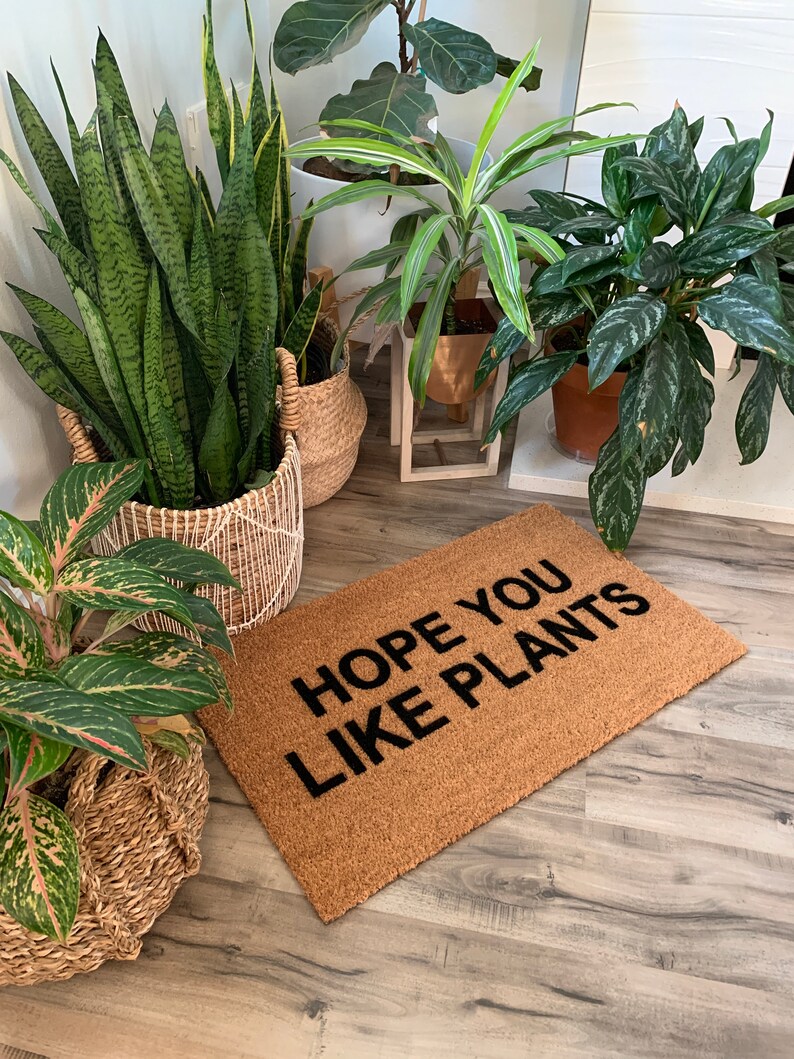 K&ouml;nnte beinhalten: Eine braune Kokosmatte mit dem Text "HOPE YOU LIKE PLANTS" in schwarzen Buchstaben. Die Fu&szlig;matte liegt vor verschiedenen Topfpflanzen.