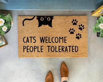 Peut inclure: Un paillasson en coco brun avec le texte "Cats Welcome People Tolerated" en lettres noires. Une silhouette de chat noir avec des yeux blancs dépasse du haut du paillasson. Il y a quatre empreintes de pattes noires sur le paillasson.