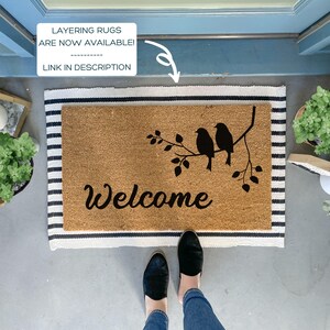 Puede incluir: Una alfombra de coco marr&oacute;n con la palabra "Welcome" en escritura negra. Tres p&aacute;jaros negros est&aacute;n posados en una rama con hojas sobre la palabra. La alfombra est&aacute; sobre una alfombra rayada blanca y negra.