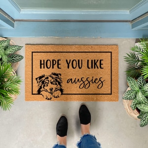 May include: A brown coir doormat with the text "HOPE YOU LIKE aussies" in black lettering. A black and white silhouette of an Australian Shepherd dog is peeking over the top of the text.