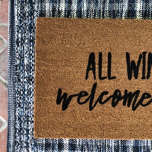 May include: A rectangular doormat with the words "ALL WELCOME" in black script. The mat is brown and surrounded by a blue and white patterned border. The mat is placed on a brick surface.