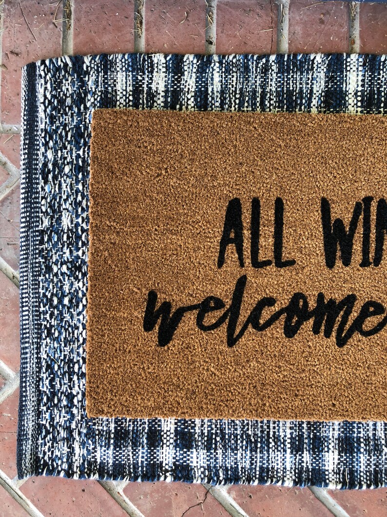 May include: A rectangular doormat with the words "ALL WELCOME" in black script. The mat is brown and surrounded by a blue and white patterned border. The mat is placed on a brick surface.
