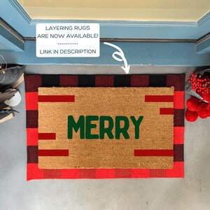 May include: A brown coir doormat with a red and black buffalo plaid border. The word "MERRY" is written in green on the doormat.