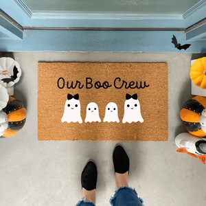 May include: A brown coir doormat with the text "Our Boo Crew" in white script. Four white ghosts with black bows are arranged in a row on the doormat.