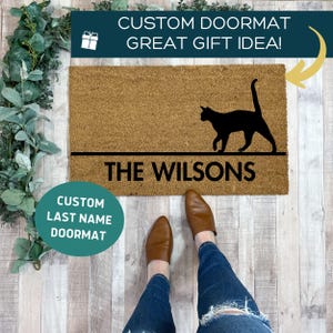 May include: A brown coir doormat with a black silhouette of a cat walking. The text "THE WILSONS" is printed in black below the cat. The text "CUSTOM LAST NAME DOORMAT" is printed in white on a teal circle.