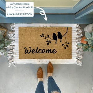 Puede incluir: Una alfombra de bienvenida de coco marr&oacute;n con la palabra "Welcome" en escritura negra y una silueta de tres p&aacute;jaros en una rama. La alfombra tiene flecos blancos alrededor de los bordes.