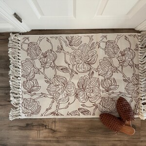 May include: A rectangular woven rug featuring a cream-colored base with a dark brown line-art floral pattern of magnolia blossoms and branches. The rug has long, knotted fringe detailing on both shorter ends and a pair of brown woven mules.