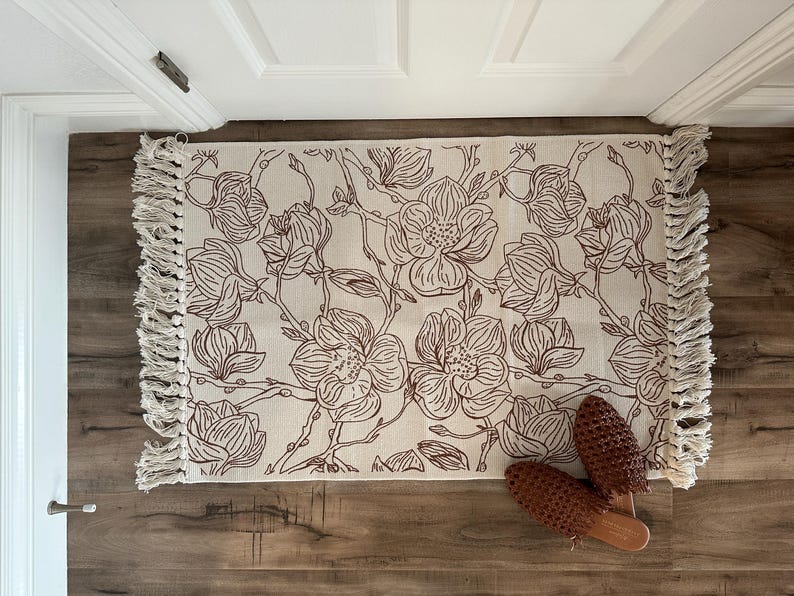 May include: A rectangular woven rug featuring a cream-colored base with a dark brown line-art floral pattern of magnolia blossoms and branches. The rug has long, knotted fringe detailing on both shorter ends and a pair of brown woven mules.
