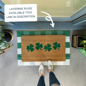 May include: A brown doormat with four green shamrock designs on it. The doormat is layered on top of a white and green checkered rug. The doormat is on a concrete surface.