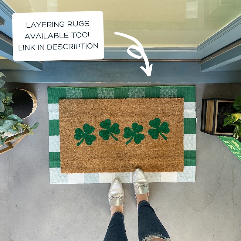 May include: A brown doormat with four green shamrock designs on it. The doormat is layered on top of a white and green checkered rug. The doormat is on a concrete surface.