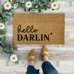 May include: A rectangular coir doormat with the words "hello DARLIN'" in black script. The mat is surrounded by faux greenery and daisies. A person is wearing yellow bow-tie flats and blue jeans.